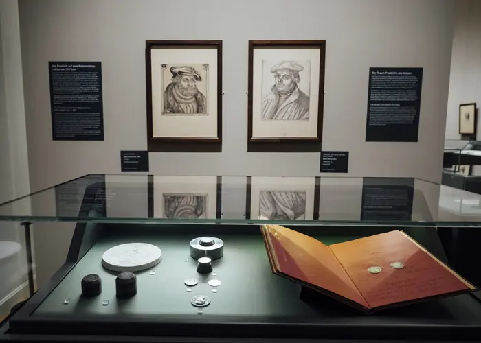 Vitrine mit einem Buch und Münzen, dahinter zwei Bilder an der Wand