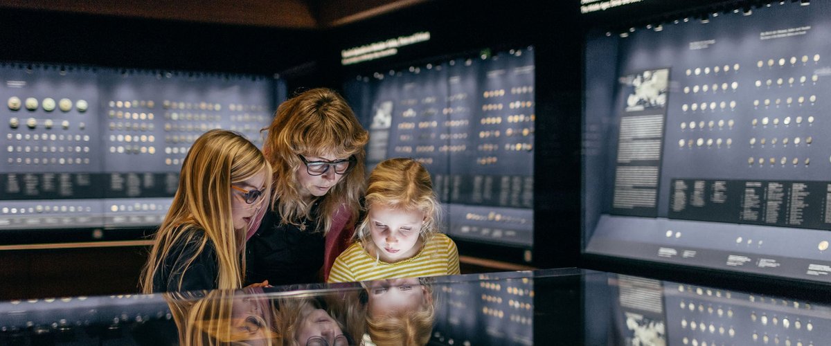 drei Besucher, darunter zwei Kinder, schauen sich Vitrine mit Münzen an